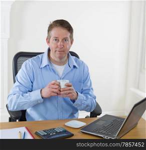 Mature man holding cup of coffee, while looking forward, with laptop, calculator, pencils, and notepad on desk. Background is white walls.