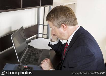 Mature man holding pen and hand against head with computer, calculator, and papers on desk