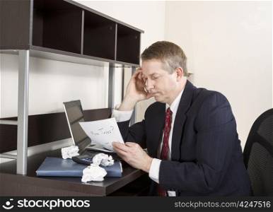 Mature man holding pen in hand against head with computer, calculator, and papers on desk