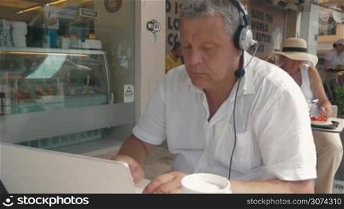 Mature man in headphones with microphone is typing in laptop and drinking coffee from paper cup.