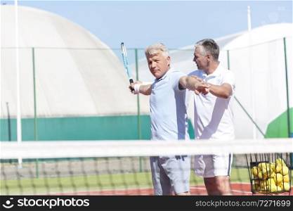 Mature man instructing friend swinging tennis racket on sunny day