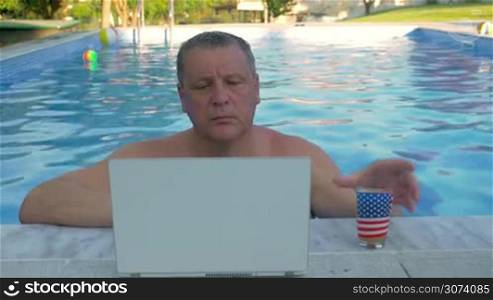 Mature man is resting by the home pool border, he is watching something interesting in laptop and drinking juice.