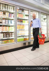 Mature man looking at mobile phone while walking in front of refrigerators in shopping centre