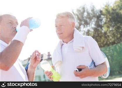 Mature man looking senior friend drinking from bottle while standing on tennis court during summer