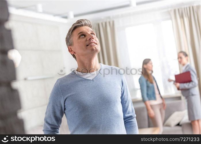 Mature man looking up while standing against saleswoman talking with woman in apartment