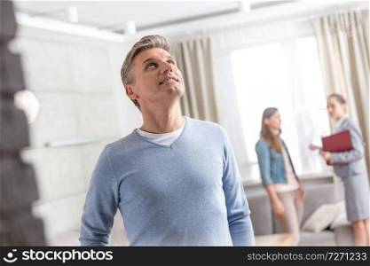 Mature man looking up while standing against saleswoman talking with woman in apartment