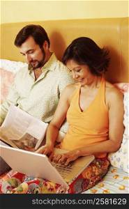 Mature man reading a newspaper with a mature woman using a laptop on the bed