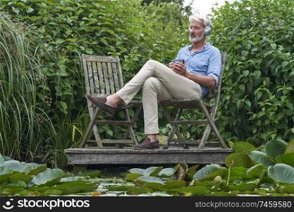 Mature Man Relaxing In Garden Listening To Music On Wireless Headphones On Jetty By Lake                             