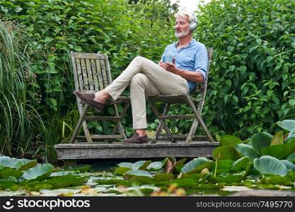 Mature Man Relaxing In Garden Listening To Music On Wireless Headphones On Jetty By Lake