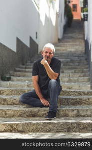 Mature man sitting on steps in the street. Senior male with white hair and beard wearing casual clothes in urban background.