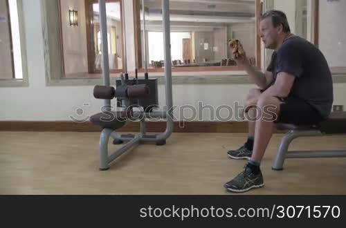 Mature man sitting on the bench in hotel gym. He relaxing after workout and wiping sweat with a towel