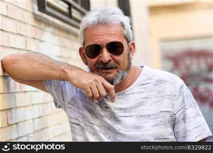 Mature man smiling at camera in urban background. Senior male with white hair and beard wearing casual clothes and aviator sunglasses.