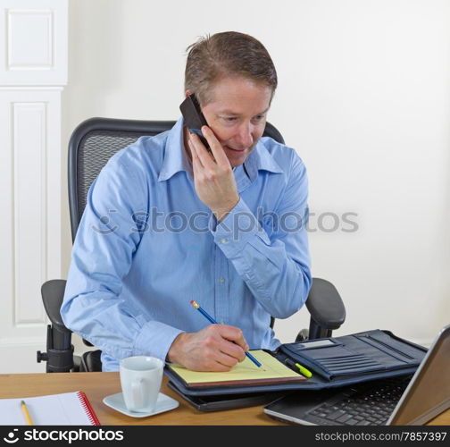 Mature man taking information via phone, pencil in hand, with laptop, calculator, coffee cup and notepad on desk. Background is white walls.