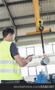 Mature manual worker with digital tablet operating crane to lift steel in industry