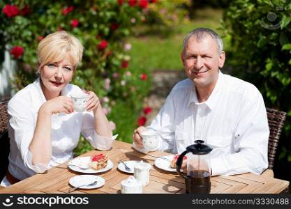 Mature or senior couple having coffee and strawberry cake on the porch in front of their home, it is summer and the roses look beautiful
