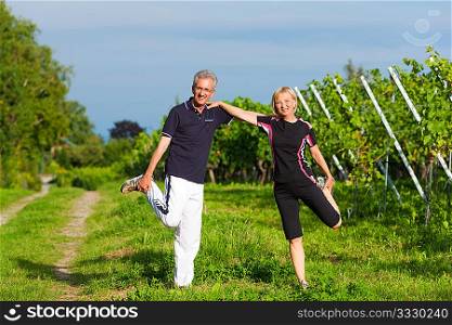 Mature or senior couple in jogging gear doing sport and physical exercise outdoors in a vineyard, stretching and gymnastics