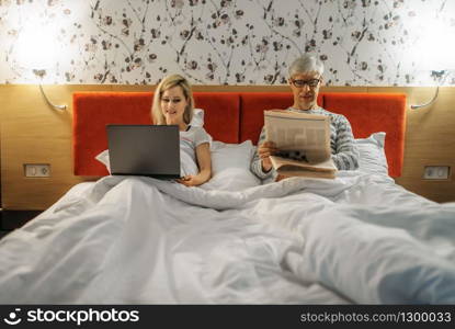 Mature wife at the laptop and husband with newspaper, couple in bedroom. Adult man and woman lying in bed before sleeping