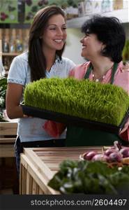 Mature woman and her daughter standing with a tray of wheatgrass in a grocery store and smiling
