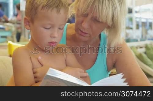 Mature woman and little boy are resting on the beach, she is reading a book to him.