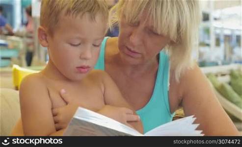 Mature woman and little boy are resting on the beach, she is reading a book to him.