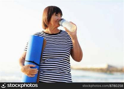 mature woman drinking water from bottle after fitness exercises or yoga on beach by sea. fitness and healthy lifestyle concept - thirsty senior woman with mat maintains water balance after sport.. mature woman drinking water from bottle after fitness exercises or yoga on beach by sea. fitness and healthy lifestyle concept - thirsty senior woman with mat maintains water balance after sport