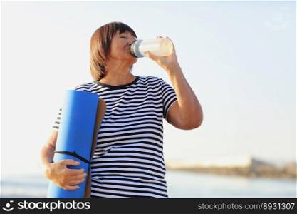 mature woman drinking water from bottle after fitness exercises or yoga on beach by sea. fitness and healthy lifestyle concept - thirsty senior woman with mat maintains water balance after sport.. mature woman drinking water from bottle after fitness exercises or yoga on beach by sea. fitness and healthy lifestyle concept - thirsty senior woman with mat maintains water balance after sport