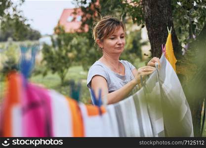 Mature woman hanging a freshly laundered bed linen on clothesline stretched between two trees in a orchard. Candid people, real moments, authentic situations