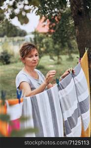 Mature woman hanging a freshly laundered bed linen on clothesline stretched between two trees in a orchard. Candid people, real moments, authentic situations