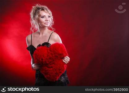 Mature woman hug big red heart . Woman mid aged blonde female wearing black evening dress holding big pillow in form of heart love symbol studio shot on red. Valentines day happiness concept
