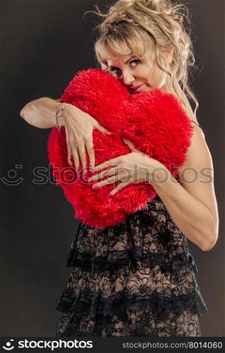 Mature woman hug big red heart . Woman mid aged blonde female wearing black evening dress holding big pillow in form of heart making love symbol from her hands studio shot on black. Valentines day happiness concept