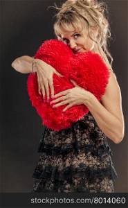 Mature woman hug big red heart . Woman mid aged blonde female wearing black evening dress holding big pillow in form of heart making love symbol from her hands studio shot on black. Valentines day happiness concept