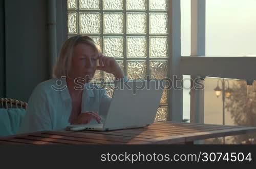 Mature woman is sitting in front of laptop on the balcony of her house.