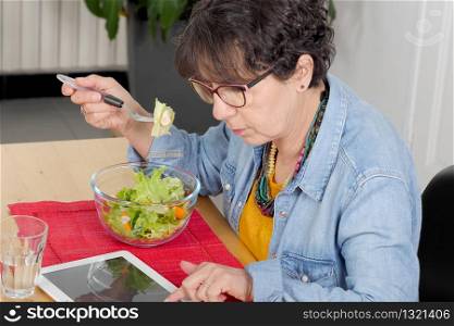 mature woman looking digital tablet while eating a salad