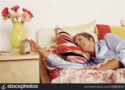 Mature woman lying in bed and reaching for an alarm clock