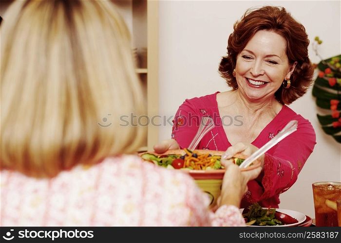 Mature woman passing a bowl of salad to her friend and smiling
