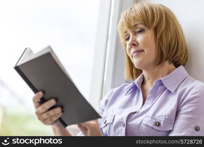 Mature woman reading book by window at home