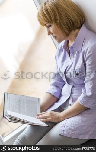 Mature woman reading book while sitting by window at home