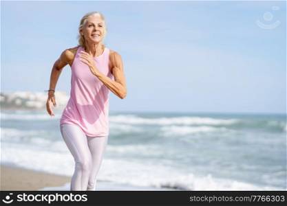 Mature woman running along the shore of the beach. Older female doing sport to keep fit. Concept of healthy living in the elderly. Senior woman in fitness clothing running along beach. Mature woman running along the shore of the beach. Older female doing sport to keep fit