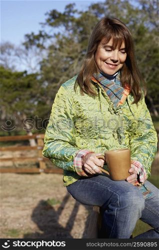 Mature woman sitting on a fence and holding a mug
