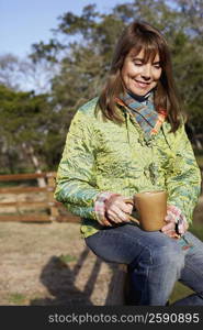 Mature woman sitting on a fence and holding a mug