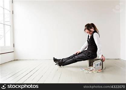 Mature woman sitting on safe in empty room, safe door open with money spilling out