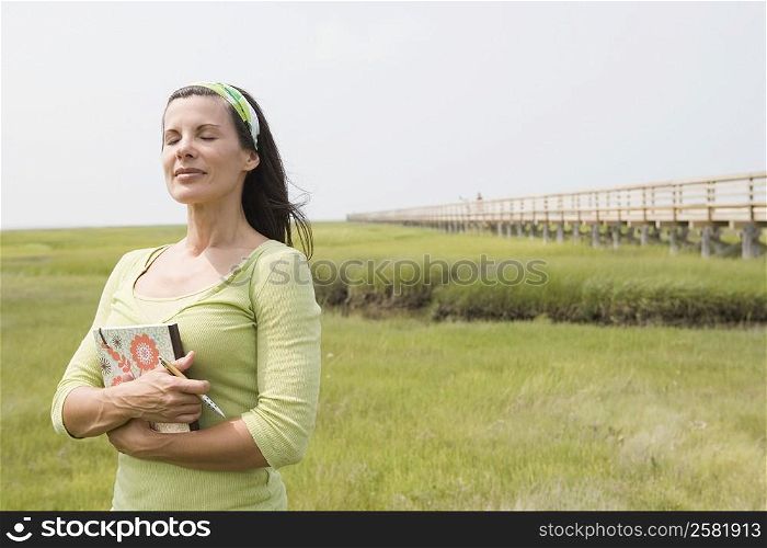 Mature woman standing in a field with her eyes closed and holding a book
