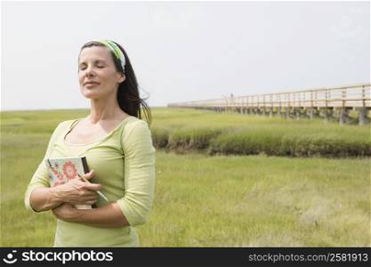 Mature woman standing in a field with her eyes closed and holding a book