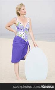 Mature woman standing on the beach with her hand on her hip and holding a body board