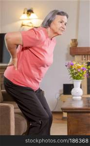 Mature woman suffering from backache at home. Massaging lower back with hand, feeling exhausted, standing in living room.