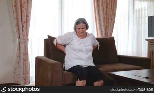 Mature woman suffering from backache at home. Massaging lower back with hands, feeling exhausted, standing in living room. Slow motion