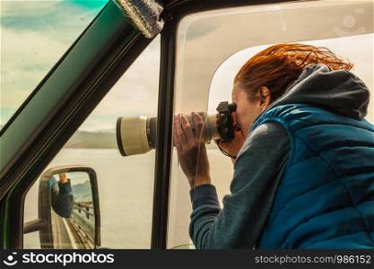 Mature woman tourist professional photographer taking photo from camper car with camera, driving on road trip. Female passenger taking picture out of window, windy weather. Photographer taking photo from camper car