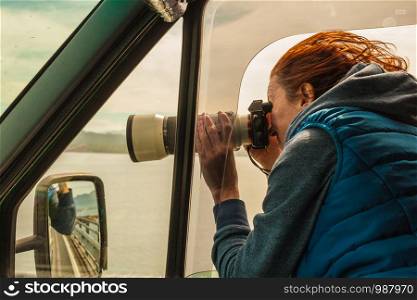 Mature woman tourist professional photographer taking photo from camper car with camera, driving on road trip. Female passenger taking picture out of window, windy weather. Photographer taking photo from camper car