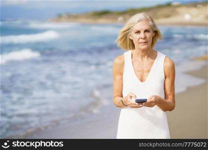 Mature woman walking on the beach using a smartphone. Elderly female enjoying her retirement at a seaside location.. Mature woman walking on the beach using a smartphone.