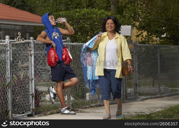 Mature woman walking with a teenage boy leaning on a chain-link fence and drinking water beside her